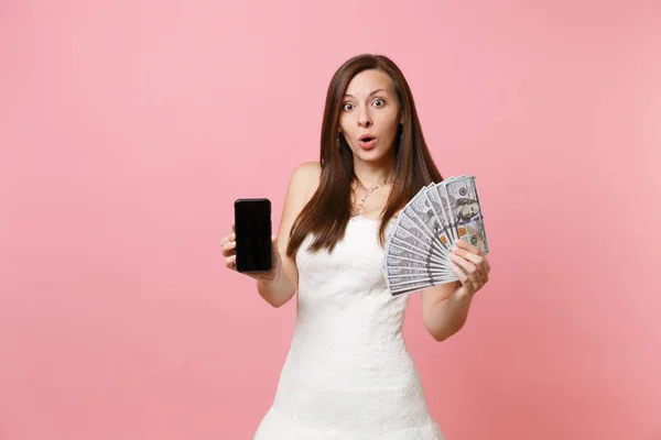 ショックを受けた花嫁バンドルたくさんのドル現金お金 ピンクの背景に分離された空白の黒い空の画面で携帯電話を保持しているウェディング ドレスの女性 結婚式の祭典の組織 コピー スペース — ストック写真