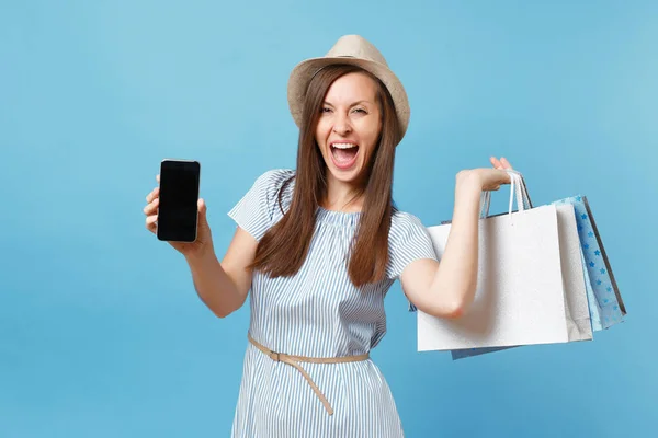 Portrait of woman in summer dress, straw hat holding packages bags with purchases after shopping, mobile cellphone with empty screen isolated on blue pastel background. Copy space for advertisement