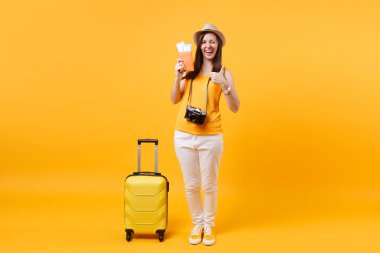 Turist kadın yaz rahat kıyafetler, pasaport ve bilet, ile şapka sarı turuncu arka plan üzerinde izole bavul. Hafta sonu kaçış üzerinde seyahat için yurt dişi. Hava uçuş yolculuk kavramı