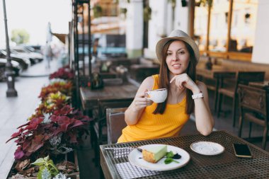 Açık havada sokak kafe Cafe kapuçino, bardak ile sarı kıyafet şapka masada oturan kadın kek, restoranda serbest zaman sırasında rahatlatıcı. Mobil ofis yazında. Yaşam tarzı dinlenme kavramı
