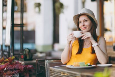 Açık havada sokak kafe Cafe kapuçino, bardak ile sarı kıyafet şapka masada oturan kadın kek, restoranda serbest zaman sırasında rahatlatıcı. Mobil ofis yazında. Yaşam tarzı dinlenme kavramı