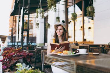 Kadın şapka, masada oturan açık havada sokak kafe Cafe fincan cappuccino ile kitap okuma, kek, restoranda serbest zaman sırasında rahatlatıcı. Mobil ofis yazında. Yaşam tarzı dinlenme kavramı