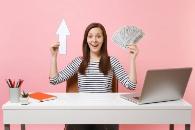 Yukarı ok, dolar nakit para çok iş pastel pembe arka plan üzerinde izole çağdaş pc dizüstü bilgisayar ile beyaz masa başında oturup paket tutan kadın hayran. Başarı iş kariyer kavramı. Kopya alanı