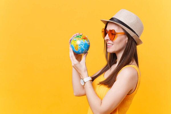 Portrait of tourist woman in summer hat, orange glasses hold in hands globe isolated on yellow background. Traveling abroad to travel weekends getaway. Air flight journey concept. Stop nature garbage