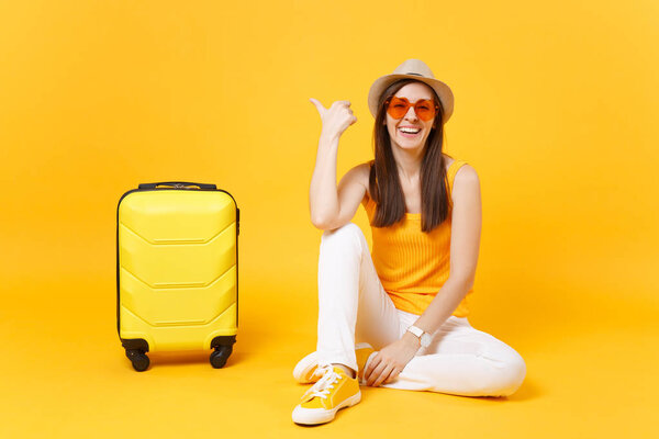 Traveler tourist woman in summer casual clothes, hat sit with suitcase isolated on yellow orange background. Female passenger traveling abroad to travel on weekends getaway. Air flight concept