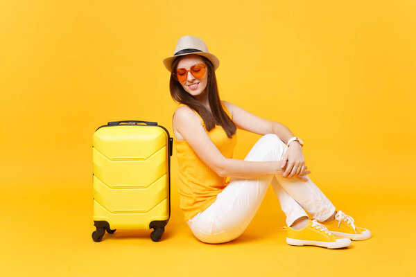 Traveler tourist woman in summer casual clothes, hat sit with suitcase isolated on yellow orange background. Female passenger traveling abroad to travel on weekends getaway. Air journey concept