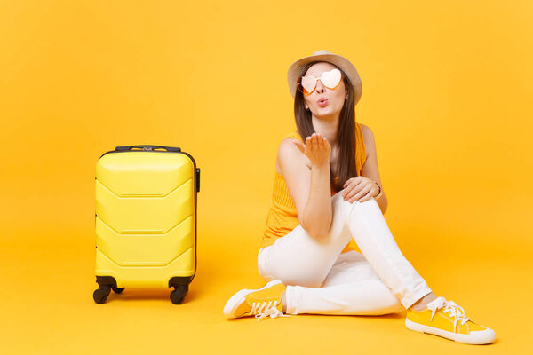Traveler tourist woman in summer casual clothes, hat sit with suitcase isolated on yellow orange background. Female passenger traveling abroad to travel on weekends getaway. Air journey concept