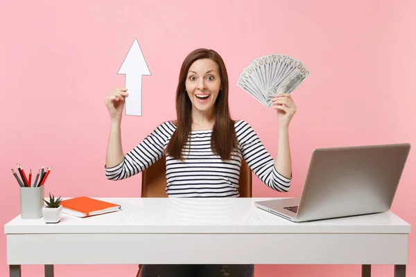 Amazed woman holding up arrow, bundle lots of dollars cash money sit work at white desk with contemporary pc laptop isolated on pastel pink background. Achievement business career concept. Copy space