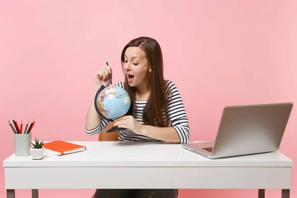 Curious woman pointing on globe with pencil, planning vacation while sit and work at white desk with contemporary pc laptop isolated on pastel pink background. Achievement business career. Copy space