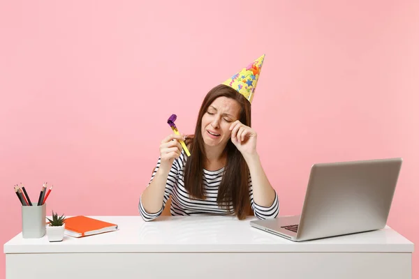 Crying woman in birthday party hat with playing pipe celebrating alone wiping tears while sit work at white desk with pc laptop isolated on pink background. Achievement business career. Copy space