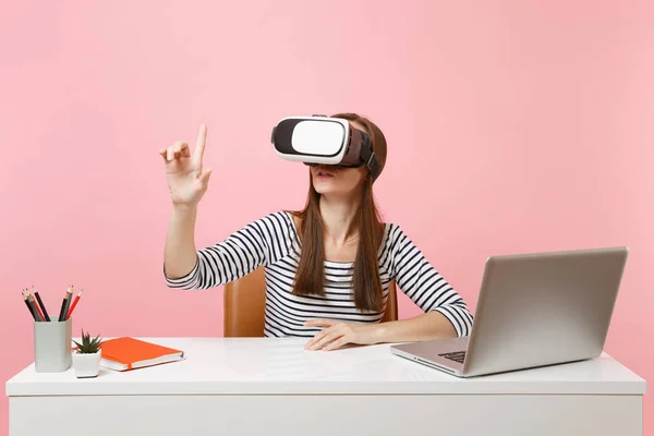 Girl in headset of virtual reality on head touch something like push on button or pointing at floating virtual screen work at desk with laptop isolated on pink background. Achievement business career