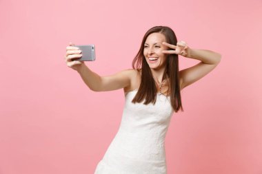 Neşeli gelin alma selfie cep telefonu pastel pembe arka plan üzerinde izole atış yapıyor zafer işaret gösteren zarif beyaz gelinlik kadın portresi. Düğün kutlama kavramı. Kopya alanı