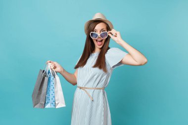 Moda çekici mutlu kadın portre yazlık elbise, hasır şapka, güneş gözlüğü izole mavi pastel arka plan üzerinde alışveriş sonra paketleri çanta satın almalarla tutarak. Kopya alanı reklam için