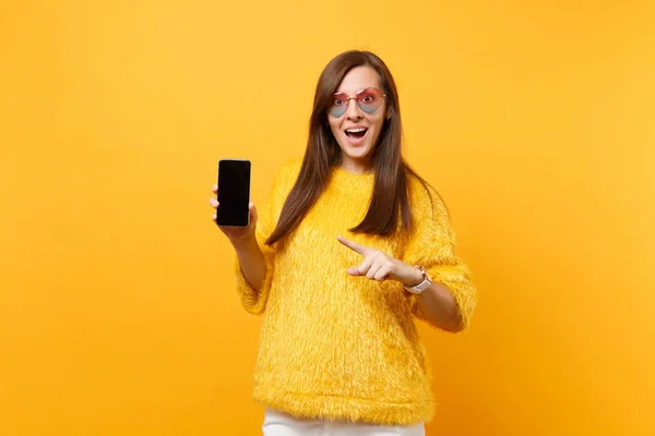 兴奋的年轻妇女在心脏眼镜指向食指在移动电话与空白黑空白屏幕被隔绝在明亮的黄色背景 人们真挚的情感 生活方式 广告专区 — 图库照片