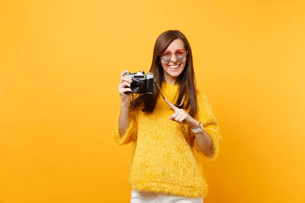 Portrait of smiling young woman in heart glasses pointing index finger on retro vintage photo camera isolated on bright yellow background. People sincere emotions, lifestyle concept. Advertising area