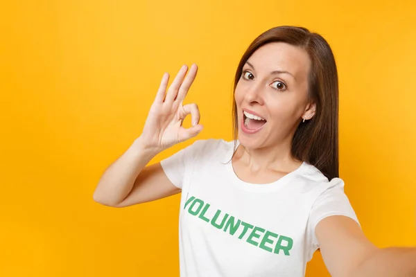 Selfie image of happy smiling satisfied woman in white t-shirt with written inscription green title volunteer isolated on yellow background. Voluntary free assistance help, charity grace work concept
