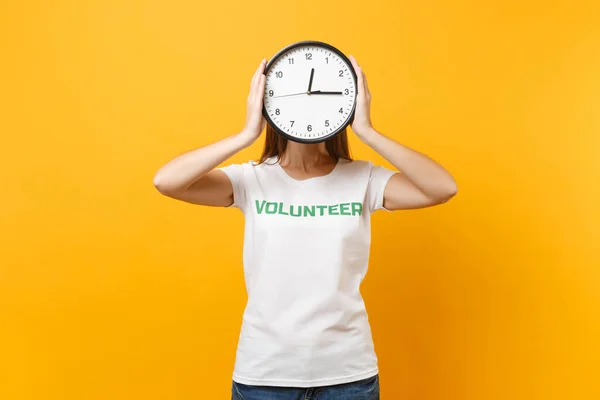 Portrait of woman in white t-shirt with written inscription green title volunteer hold round clock isolated on yellow background. Voluntary free assistance help, charity grace work time concept.