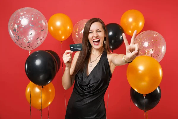 快乐的年轻女孩穿着黑色礼服尖叫着持有信用卡和显示胜利标志红色背景气球 国际妇女节 新年快乐 生日样机节日派对理念 — 图库照片
