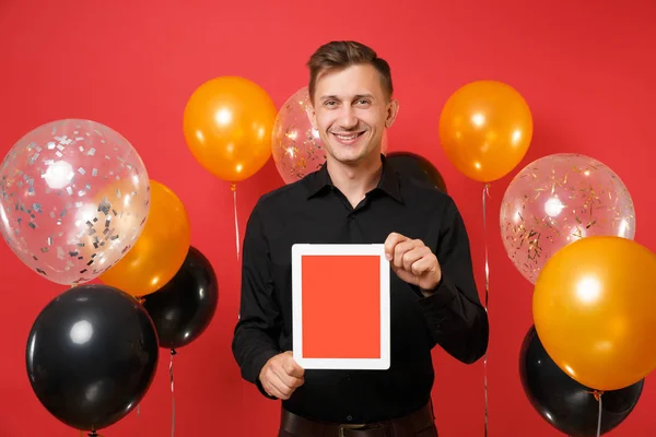 Smiling young man in black classic shirt hold tablet pc computer with blank black empty screen on bright red background air balloons. Women\'s Day, Happy New Year birthday mockup holiday party concept