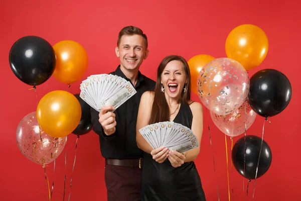 Couple in black clothes celebrating birthday holiday party, hold cash money isolated on bright red background air balloons. St. Valentine International Women Day Happy New Year 2019 concept. Mock up