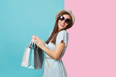 Moda çekici mutlu kadın portre yazlık elbise, hasır şapka, güneş gözlüğü izole mavi pastel arka plan üzerinde alışveriş sonra paketleri çanta satın almalarla tutarak. Kopya alanı reklam için
