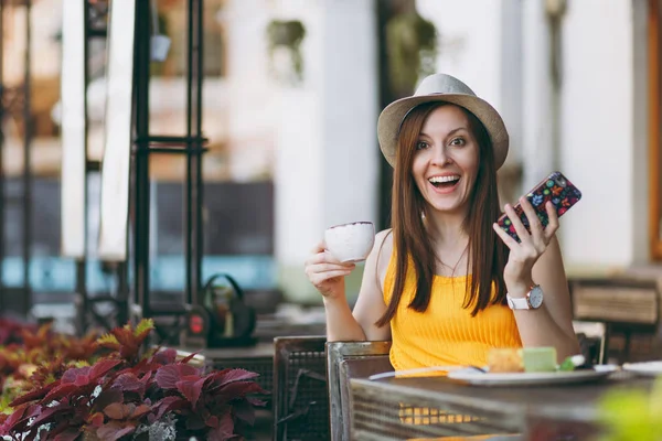 Woman in outdoors street coffee shop cafe sitting at table in hat with cup of cappuccino cake, using mobile phone, relaxing in restaurant on free time. Mobile Office in summer. Lifestyle rest concept