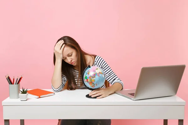 Tired woman with closed eyes lean on hand hold globe having problems with vacation planning while sit, work at office with laptop isolated on pink background. Achievement business career. Copy space