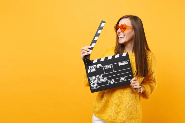 Laughing young woman in orange heart eyeglasses looking aside and holding classic black film making clapperboard isolated on yellow background. People sincere emotions, lifestyle. Advertising area