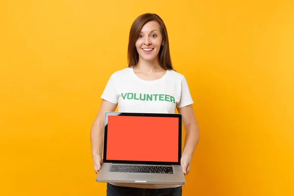 Woman in white t-shirt written inscription green title volunteer hold laptop pc computer, blank empty screen isolated on yellow background. Voluntary free assistance help, charity grace work concept