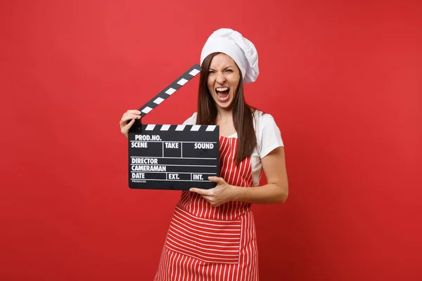 Housewife female chef cook or baker in striped apron, white t-shirt, toque chefs hat isolated on red wall background. Woman holding classic black film making clapperboard. Mock up copy space concept