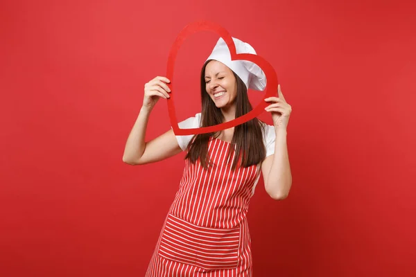 Housewife female chef cook or baker in striped apron, white t-shirt, toque chefs hat isolated on red wall background. Smiling housekeeper woman holding wooden red heart. Mock up copy space concept