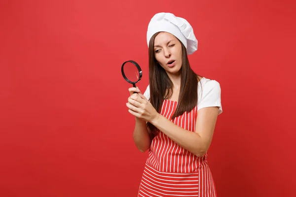 Housewife female chef cook or baker in striped apron white t-shirt toque chefs hat isolated on red wall background. Housekeeper woman hold look through magnifying glass. Mock up copy space concept