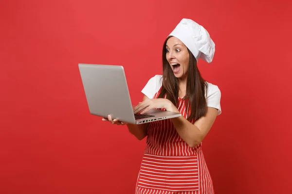 Housewife female chef cook or baker in striped apron, white t-shirt, toque chefs hat isolated on red wall background. Fun housekeeper woman looking for recipe in laptop pc. Mock up copy space concept