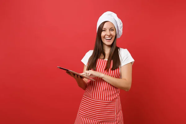Housewife female chef cook or baker in striped apron, white t-shirt, toque chefs hat isolated on red wall background. Fun housekeeper woman looking for recipe in tablet pc. Mock up copy space concept