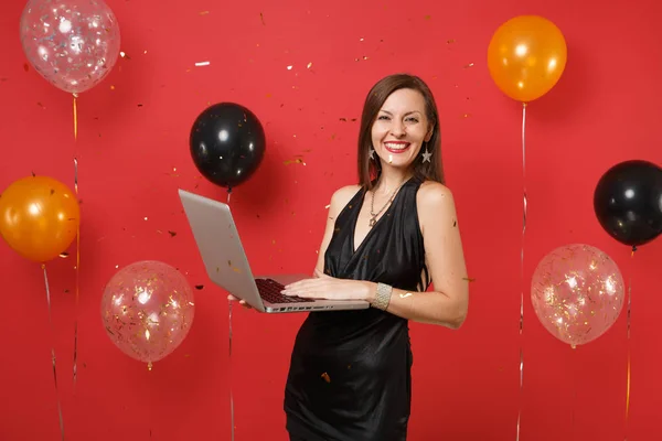 Joyful young girl in little black dress working on laptop pc computer while celebrating on red background air balloons. International Women\'s Day, Happy New Year birthday mockup holiday party concept
