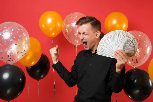 Happy man in classic shirt screaming clenching fist like winner holding bundle lots of dollars, cash money in hand on red background air balloons. Happy New Year birthday mockup holiday party concept