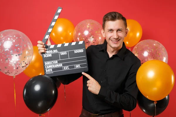 Handsome young man in black shirt pointing index finger on classic black film making clapperboard in hands on bright red background air balloons. Happy New Year, birthday mockup holiday party concept