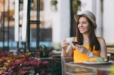 Açık havada sokak kafe Cafe cappucino kek, bardak ile şapkalı masada oturan istimal hareket eden telefon, ücretsiz zamanında restoranda rahatlatıcı kadın. Mobil ofis yazında. Yaşam tarzı dinlenme kavramı