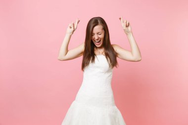 Portrait of happy bride woman in white wedding dress waiting for special moment, keeping fingers crossed, eyes closed isolated on pastel pink background. Wedding concept. Copy space for advertisement clipart