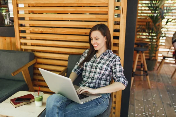 Woman in outdoors street summer coffee shop wooden cafe sitting in casual clothes, working on modern laptop pc computer, relaxing during free time. Mobile Office. Lifestyle freelance business concept