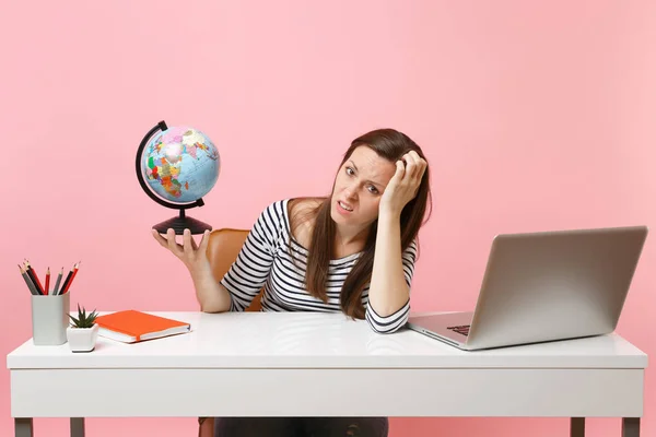 Irritated woman leaning on hand holding globe having problems with vacation planning while sit, work at office with laptop isolated on pastel pink background. Achievement business career. Copy space
