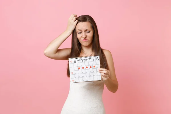 Puzzled guilty bride woman in wedding dress clinging in head, looking on female periods calendar for checking menstruation days isolated on pink background. Medical, healthcare, gynecological concept