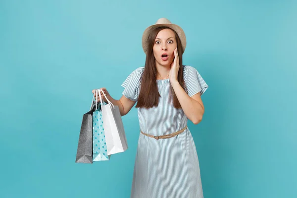 Portrait pretty shocked sad beautiful caucasian woman in summer dress, straw hat holding packages bags with purchases after shopping isolated on blue pastel background. Copy space for advertisement