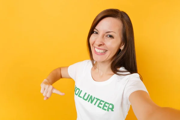 Selfie image of happy smiling satisfied woman in white t-shirt with written inscription green title volunteer isolated on yellow background. Voluntary free assistance help, charity grace work concept