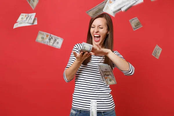 Crazy woman in striped clothes screaming scattering throwing out money banknotes lots of dollars isolated on bright red wall background. People sincere emotions, lifestyle concept. Mock up copy space