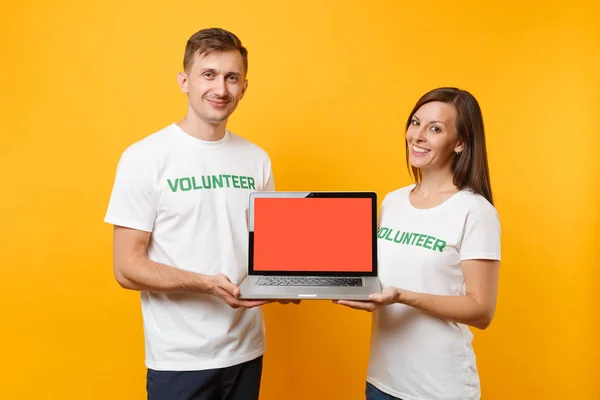 Colleagues couple in white t-shirt inscription volunteer hold laptop pc computer, blank empty screen isolated on yellow background. Voluntary free work, assistance help charity grace teamwork concept