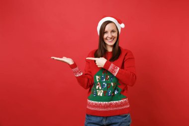 Örme kazak, Noel şapka işaret eden bir ele, işaret parmağı bir kenara izole kırmızı duvar arka plan üzerinde gülümseyen genç Noel Baba kız. Mutlu yeni yıl 2019 kutlama tatil parti kavramı. Kopyalama alanı alay