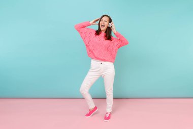 Tam uzunlukta örme gül kazak, beyaz pantolon, parlak pembe mavi pastel duvar arka planda Studio izole kulaklık mutlu genç kadın portresi. Moda yaşam tarzı kavramı. Kopyalama alanı alay