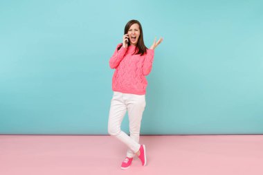 Tam uzunlukta örme gül kazaklı kadın portresi, cep telefonu parlak pembe mavi pastel duvar arka planda Studio izole konuşurken beyaz pantolon. Moda yaşam tarzı kavramı. Kopyalama alanı alay