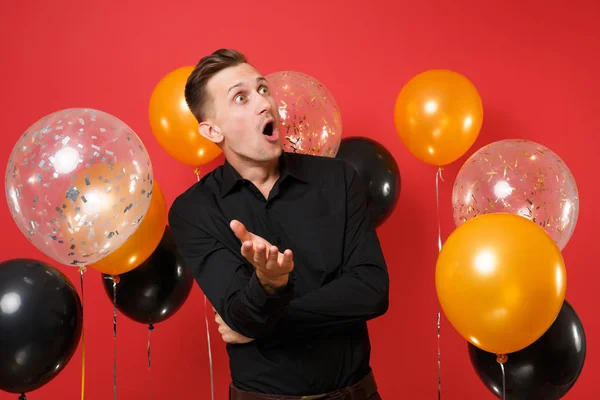 Shocked young man in black classic shirt looking up spreading hands on bright red background air balloons. Valentine's, International Women's Day, Happy New Year birthday mockup holiday party concept